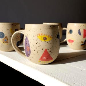 May include: A set of four speckled ceramic mugs with abstract face designs. Each mug features a unique combination of colorful shapes, including triangles, ovals, and circles, in red, yellow, blue, and purple. The mugs have a neutral beige base color.