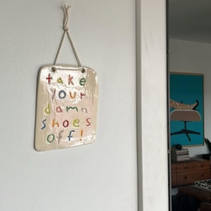 May include: A rectangular, cream-colored ceramic wall plaque with rounded corners, suspended by a light-colored rope. The plaque features the embossed, multi-colored text "take your damn shoes off!" in a playful, hand-lettered style font.