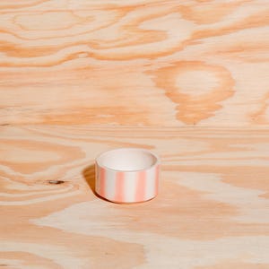 May include: A small, cylindrical ceramic bowl featuring a white interior and a exterior decorated with vertical, soft pink stripes. The bowl has a flat base and a smooth, rounded rim, presenting a simple, open-top design for holding small items.