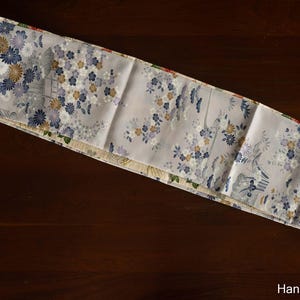 May include: A light gray fabric with a floral pattern in blue, gold, and white. The fabric is folded and lies on a dark brown surface. The text "Hanami Clothing" is visible.