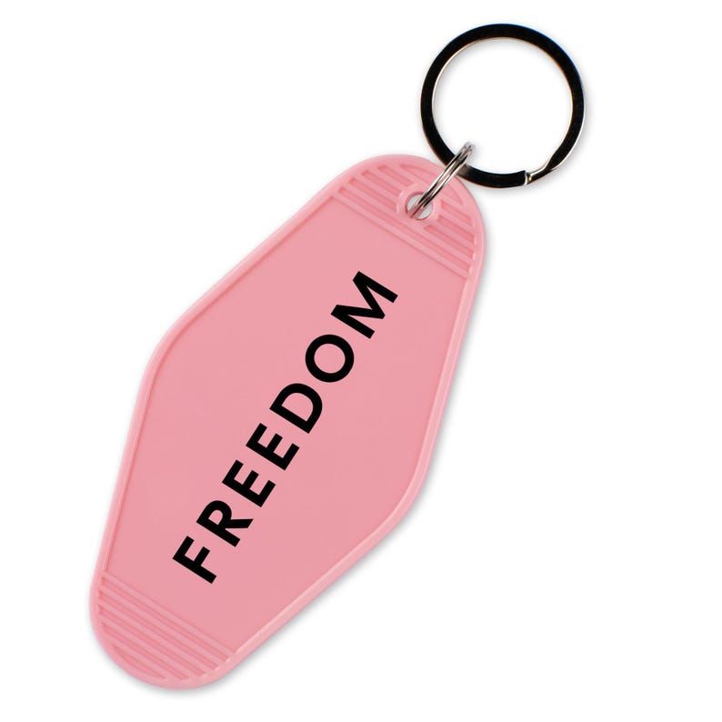 May include: A pink motel-style keychain with the word "FREEDOM" printed in black. The keychain has a ridged design at the top and bottom and a black metal keyring.