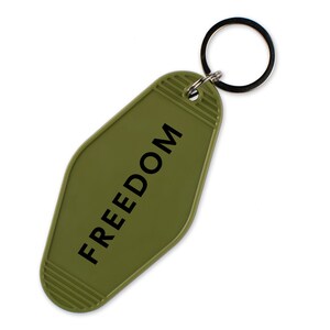 May include: Olive green keychain with the word "FREEDOM" printed in black. The keychain has a retro motel style design with a metal key ring.