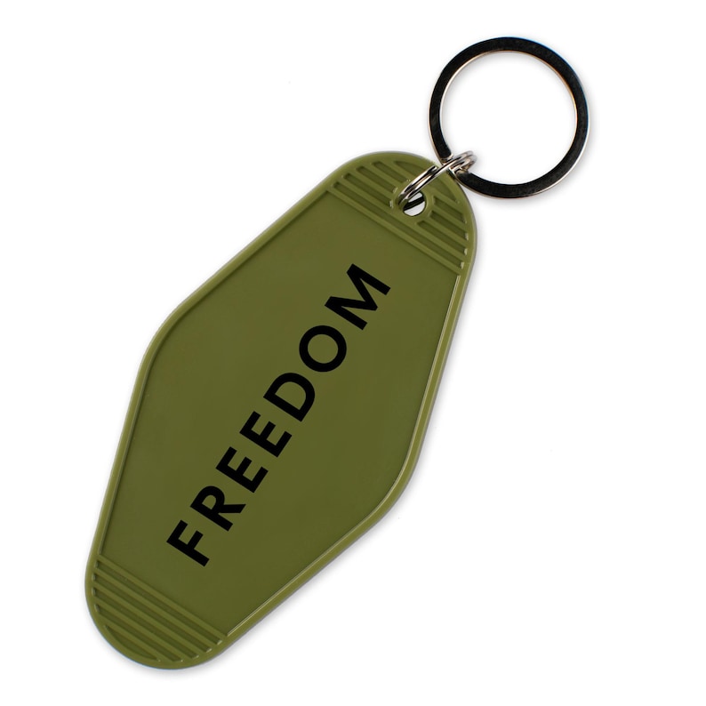 May include: Olive green keychain with the word "FREEDOM" printed in black. The keychain has a retro motel style design with a metal key ring.