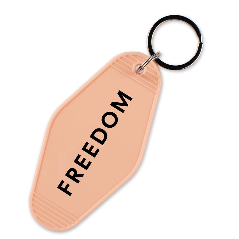 May include: A peach-colored motel-style keychain with the word "FREEDOM" printed in black. The keychain has a metal ring attached for keys. The design is simple and retro, with a textured bottom.