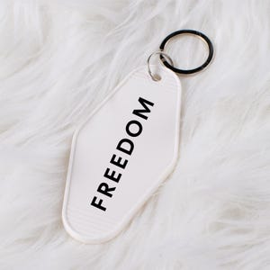 May include: A white motel-style keychain with the word "FREEDOM" printed in black. The keychain has a silver key ring attached and is set against a fluffy white background.