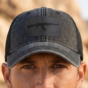 May include: A charcoal grey distressed baseball cap featuring a black rectangular patch on the front panel. The patch displays a black silhouette of an AK-style rifle. The cap has a structured front and a mesh back with a curved brim.