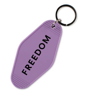 May include: A lavender motel-style keychain with the word "FREEDOM" printed in black. The keychain has a silver key ring attached at the top. The background is white.