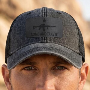 May include: A charcoal gray distressed baseball cap with a black mesh back and a rectangular patch on the front. The patch features a black star, a rifle silhouette, and the text COME AND TAKE IT printed in a bold, sans-serif font.