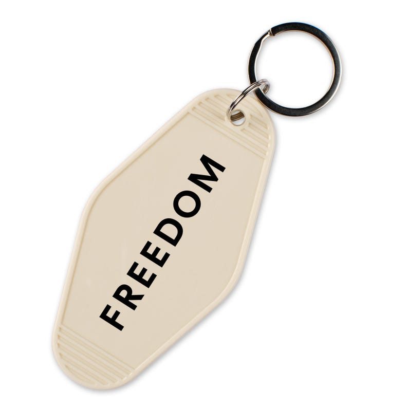 May include: Cream-colored motel-style keychain with the word "FREEDOM" printed in black. The keychain has a rectangular shape with rounded edges and a silver key ring attached at the top.