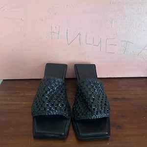 Op de afbeelding: Een paar zwarte geweven instapsandalen met vierkante tenen. De sandalen hebben een geweven ontwerp op de bovenste bandjes. De achtergrond is een roze muur met het woord "нищет" erop geschreven.