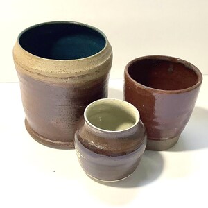 May include: Three handcrafted ceramic vessels in varying sizes. The largest is a tall, cylindrical pot with a dark brown exterior and a teal interior. The other two are smaller, with a reddish-brown and a purple-brown color scheme. The pottery has a rustic, earthy aesthetic.