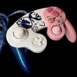 Könnte beinhalten: Ein individueller Videospiel-Controller mit weißem Gehäuse und rosa Akzenten. Der Controller verfügt über ein blau-grünes geflochtenes Kabel. Die Oberseite des Controllers zeigt eine Grafik einer Person und eines Pferdes.