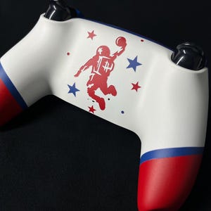 May include: A white video game controller featuring a red graphic of an astronaut jumping with a ball. The handles have red and blue bands. The design includes small red and blue stars surrounding the astronaut figure on the central white surface.