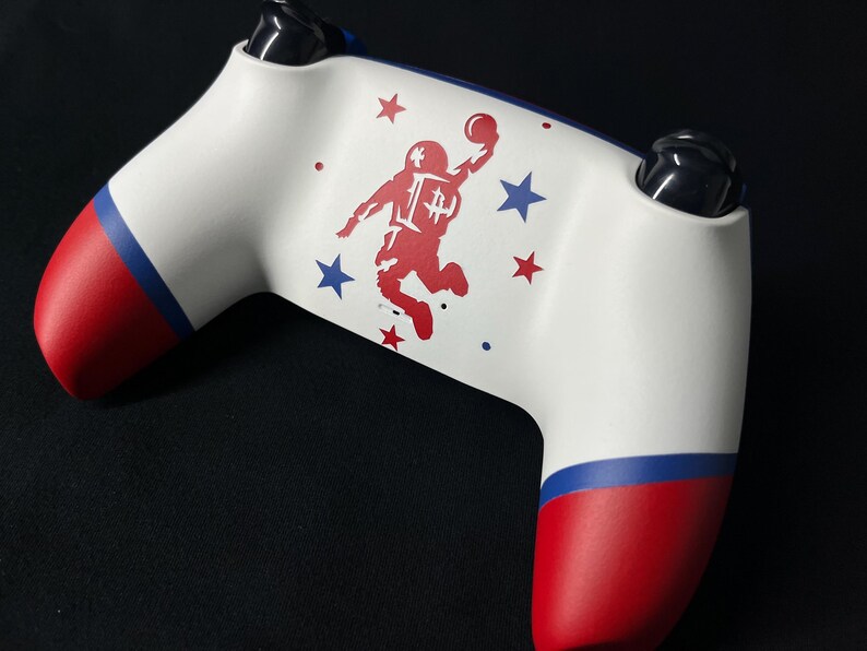 May include: A white video game controller featuring a red graphic of an astronaut jumping with a ball. The handles have red and blue bands. The design includes small red and blue stars surrounding the astronaut figure on the central white surface.