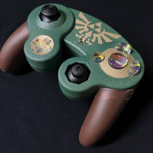 May include: A custom video game controller featuring a textured dark green shell with brown hand grips. The face displays a gold Triforce emblem, black analog sticks, and iridescent, multi-colored action buttons and a directional pad.