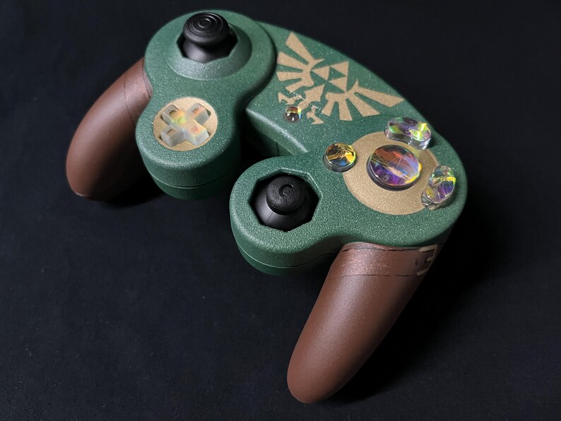 May include: A custom video game controller featuring a textured dark green shell with brown hand grips. The face displays a gold Triforce emblem, black analog sticks, and iridescent, multi-colored action buttons and a directional pad.