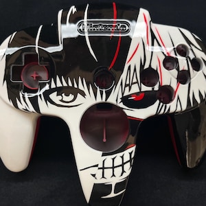 May include: A Nintendo 64 video game controller featuring a custom black and white graphic design. The controller displays a split-face illustration with red accents, a central circular port, a directional pad, and four circular action buttons.