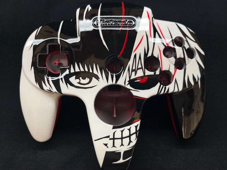May include: A Nintendo 64 video game controller featuring a custom black and white graphic design. The controller displays a split-face illustration with red accents, a central circular port, a directional pad, and four circular action buttons.