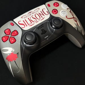 May include: A custom game controller featuring a grey and black finish with red accents. The center touchpad displays the text Hollow Knight Silksong in red and black. A graphic of a white and red character is printed on the left handle.