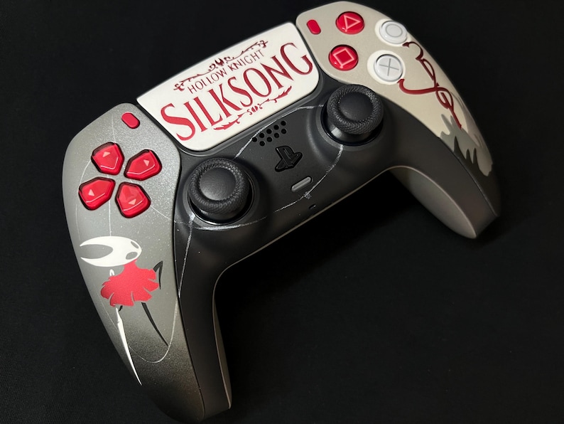 May include: A custom game controller featuring a grey and black finish with red accents. The center touchpad displays the text Hollow Knight Silksong in red and black. A graphic of a white and red character is printed on the left handle.