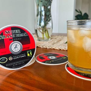 May include: A stack of coasters with a red and black design featuring the text "Super Smash Bros. Melee". A glass of iced beverage sits on a matching coaster. The coasters are on a wooden table.