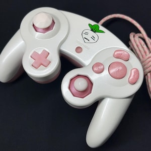 May include: A white and light pink wired video game controller featuring a small graphic of a white turnip with green leaves. The controller includes a directional pad, two analog sticks, and several action buttons in pink and white.