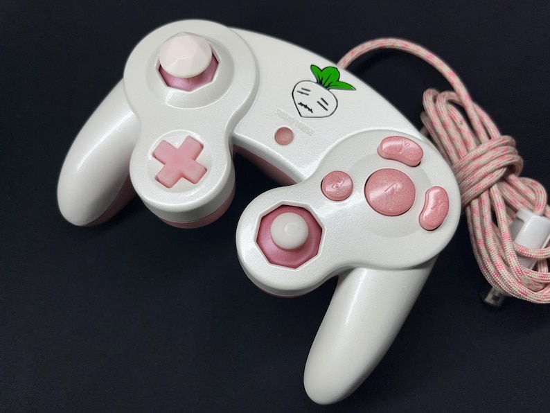 May include: A white and light pink wired video game controller featuring a small graphic of a white turnip with green leaves. The controller includes a directional pad, two analog sticks, and several action buttons in pink and white.