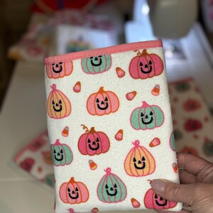 May include: A white kitchen towel with a pink and green pumpkin pattern. The pumpkins have black jack-o-lantern faces and are surrounded by candy corn.