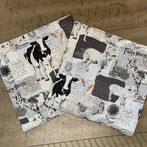 May include: Two white fabric placemats with black and white illustrations of farm animals, including cows, sheep, chickens, and a pig. The placemats feature a vintage, farmhouse style with handwritten text and a black and white color scheme. The text on the placemats reads "Farm House" and "Cuisine".