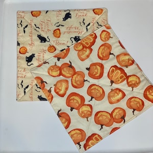 May include: Two cream-colored fabric pot holders with a Halloween theme. One pot holder has a black cat and bats with the text "Trick or Treat", "Spooktacular", "Haunted Night", and "Boo!" The other pot holder has a pattern of orange and yellow jack-o'-lanterns with the text "BOO".