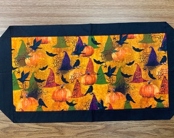 Può includere: Un runner da tavolo di Halloween nero e arancione con un motivo di cappello da strega, zucca e corvo.