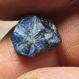 Pode incluir: Uma aproximação de uma gema de safira natural bruta. A safira é de cor azul profunda com uma superfície ligeiramente texturizada e algumas inclusões azuis e amarelas mais claras. A gema tem uma forma irregular.