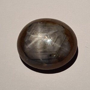 Peut inclure: Une pierre précieuse polie, de couleur brun foncé, avec un motif en forme d'étoile sur sa surface. La pierre a une forme arrondie et une finition lisse et réfléchissante. L'effet d'étoile est créé par la lumière réfléchie par les inclusions.