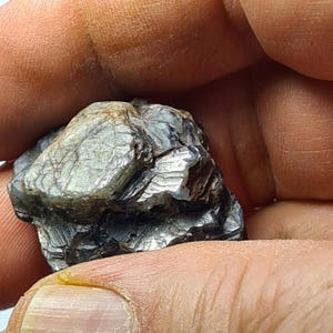 May include: A close-up of a dark, metallic mineral specimen. The stone has a layered, textured appearance with shades of gray and silver. The mineral is held in a person's hand.