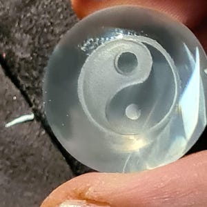 Könnte beinhalten: Eine klare, runde Glasperle mit einem mattierten Yin-und-Yang-Symbol, das in die Mitte geschnitzt ist. Die Perle wird zwischen zwei Fingern gehalten, was das komplizierte Design und die Lichtbrechung durch das Glas zeigt.