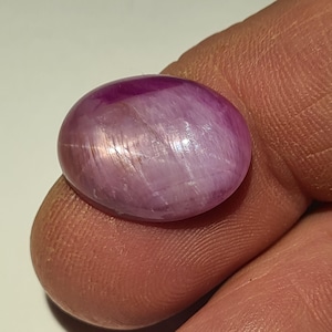 Pode incluir: Uma gema oval polida, com tons de rosa e roxo. A gema tem uma superfície lisa com linhas internas sutis e um leve brilho, sugerindo um efeito de estrela. A pedra é segurada entre os dedos, mostrando seu tamanho e cor.
