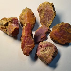 May include: A collection of rough, uncut gemstones. The stones display a range of pink and reddish-brown hues, with some areas showing a golden-yellow crust. The irregular shapes and textures of the stones suggest they are in their natural, unpolished state.