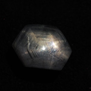 Könnte beinhalten: Ein facettierter, grauer Edelstein mit einem leicht schillernden Glanz. Der Edelstein ist in hexagonaler Form geschliffen.