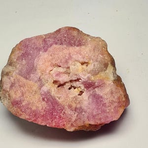 May include: A rough, uncut gemstone with a mix of pink, purple, and tan hues. The stone has a textured surface with a cavity in the center. The overall shape is irregular, showcasing natural geological formations.