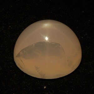 Könnte beinhalten: Ein runder, durchscheinender, hellrosa Edelstein mit einer glatten, polierten Oberfläche. Der Stein reflektiert das Licht und erzeugt einen hellen Punkt in der Mitte.
