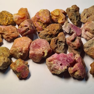 May include: A collection of rough, uncut gemstones in shades of pink, red, and brown. The stones vary in size and shape, with a textured, natural appearance. The gemstones are scattered across a white surface, showcasing their raw beauty.
