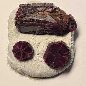 May include: A collection of raw, deep red ruby gemstones. One large, rough ruby sits atop a white, textured base, with two smaller, faceted rubies below. The gemstones display a rich, dark red color with visible internal structures.