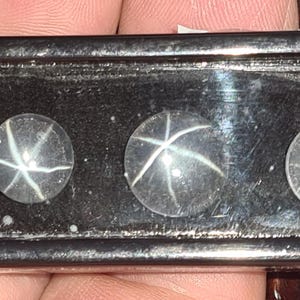 May include: Three clear, round gemstones with a star-like pattern of white lines. The gems are set on a dark, textured surface. The star effect is created by intersecting lines within the stones, creating a six-pointed star.