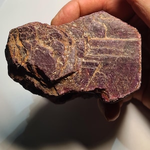 May include: A rough-cut, natural ruby gemstone with a deep red-purple hue. The stone has a textured surface with visible crystalline structures and areas of lighter, brownish tones. The ruby is held in a hand, showcasing its raw, unpolished form.