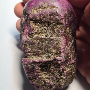 Puede incluir: Una gema de rubí natural en bruto. El rubí tiene un tono rojo rosado intenso con áreas de matriz marrón y beige. La superficie de la piedra es texturizada e irregular, revelando su estado bruto y sin pulir. La gema se sostiene en una mano.