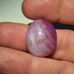 Può includere: Una gemma ovale con una miscela di tonalità rosa e lavanda. La superficie lucida riflette la luce, rivelando motivi interni e una qualità traslucida. La gemma è tenuta in una mano, evidenziandone le dimensioni e la forma.