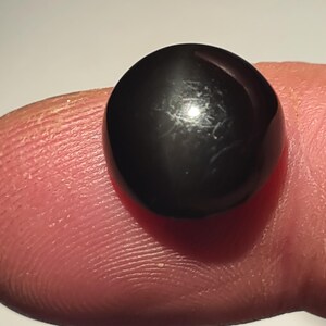 Könnte beinhalten: Ein glatter, runder, schwarzer Cabochon-Edelstein mit einer polierten, reflektierenden Oberfläche. Der Stein hat ein dunkles, undurchsichtiges Aussehen mit subtilen Lichtreflexen auf der gewölbten Oberseite und einer gleichmäßigen Kreisform.