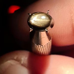 May include: A metallic ring featuring a polished, oval-shaped brown star gemstone set in a four-prong mounting. The band of the ring displays a textured, cross-hatched pattern on its surface, providing a distinct contrast to the smooth, reflective stone.