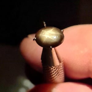 以下が含まれることがあります： 4本の爪の金属製台座にセットされた、磨き上げられた楕円形の茶色のスターサファイアの宝石。この石は、滑らかなカボションカットの表面全体に、中心から光を反射する6条の星のような効果をはっきりと示しています。