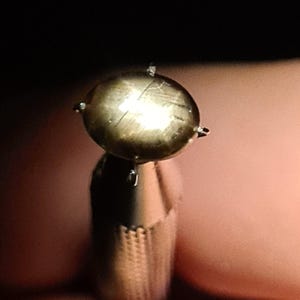 以下が含まれることがあります： 表面に星形の光の反射が見られる、丸いメタリック調のカボションカットの宝石。この石は、磨き上げられた質感のある円筒形の金属製ベースの上で、4本の爪によってしっかりと固定されています。
